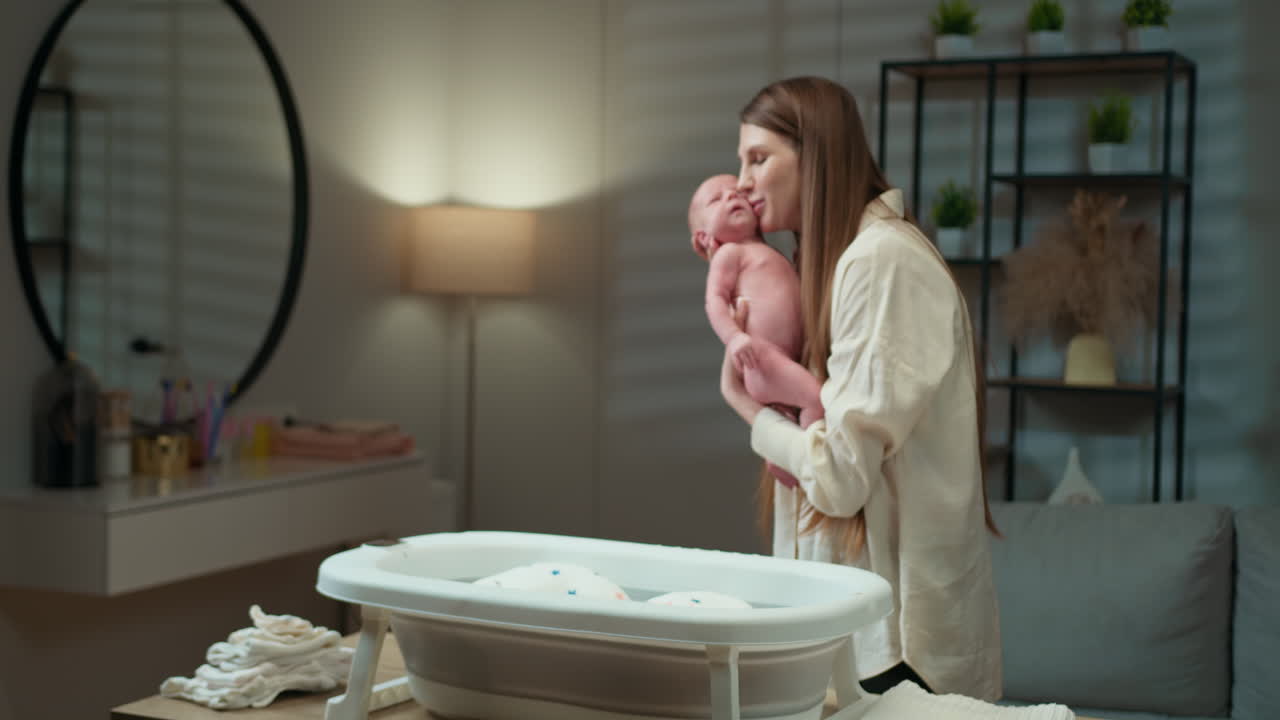 Une mère embrasse tendrement son nouveau-né tout en le tenant près d’une baignoire pour bébé dans une pièce à la lumière douce.