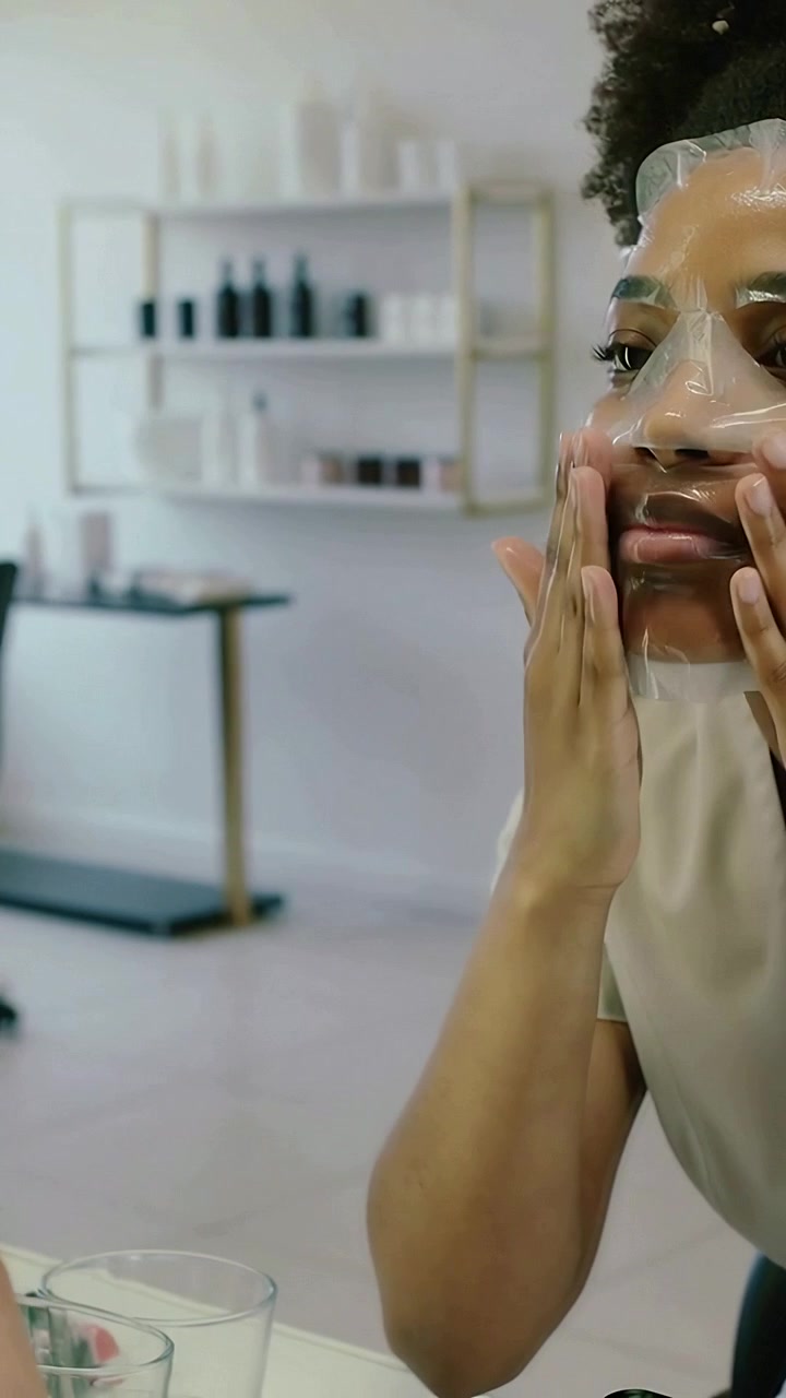 Video verticale: Donna con top chiaro che tiene una maschera in tessuto trasparente e la applica allo specchio da toeletta per la cura della pelle