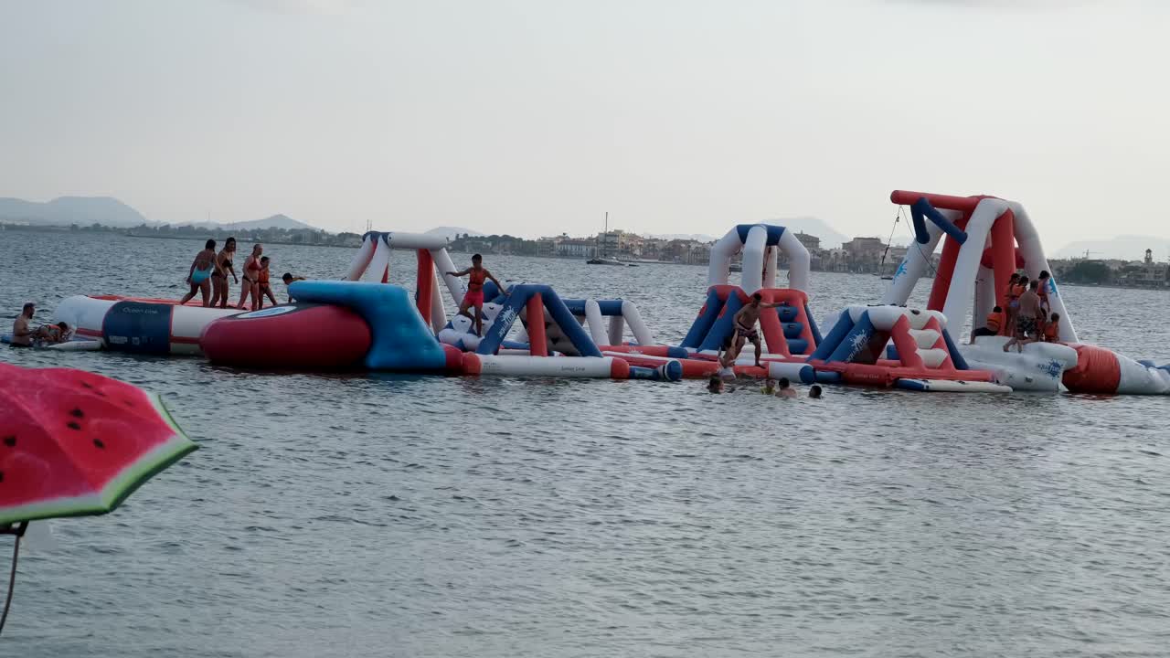 At the beach, inflatable slides float on the water, filled with excited children and adults jumping, sliding, and splashing into the waves, creating joyful chaos.