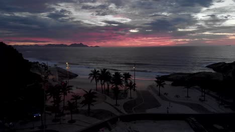 descenso aéreo y panorámica que revelan la playa de los diablos en río de janeiro con la roca arpoador en primer plano y el barrio de ipanema y un paisaje urbano más amplio en el fondo contra un cielo de color magenta
