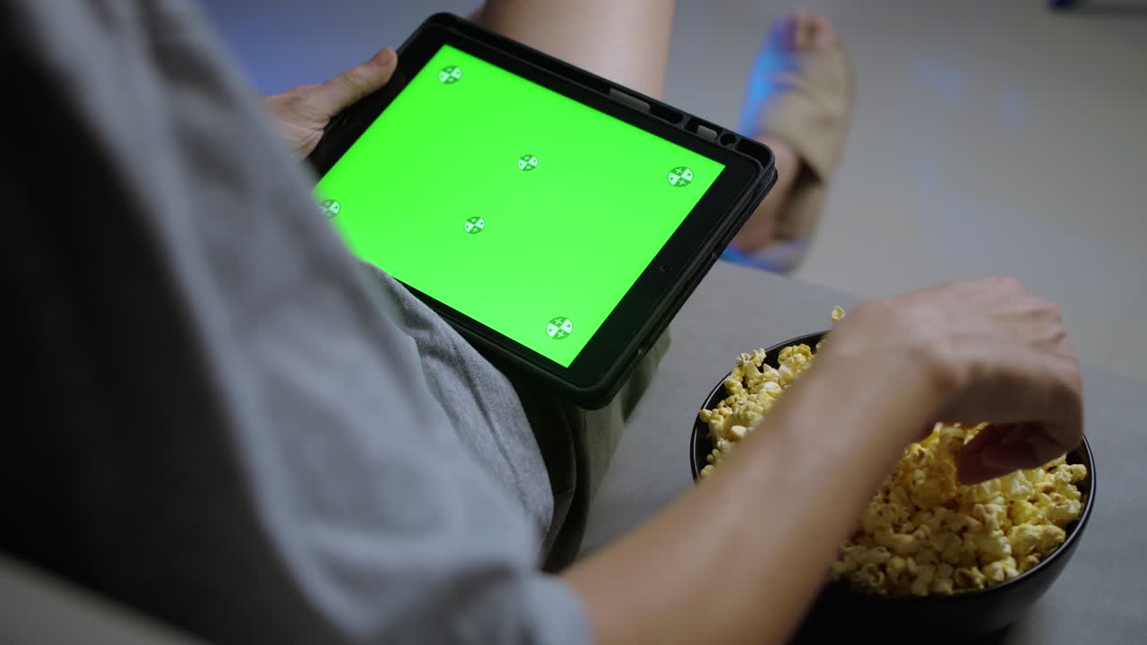 녹색 터치 스크린의 태블릿을 들고 집에서 소파에서 팝콘을 먹는 남자.