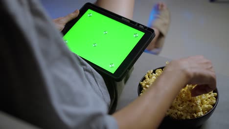 de cerca, un hombre sosteniendo una tableta con pantalla táctil verde y comiendo palomitas de maíz en el sofá en casa.