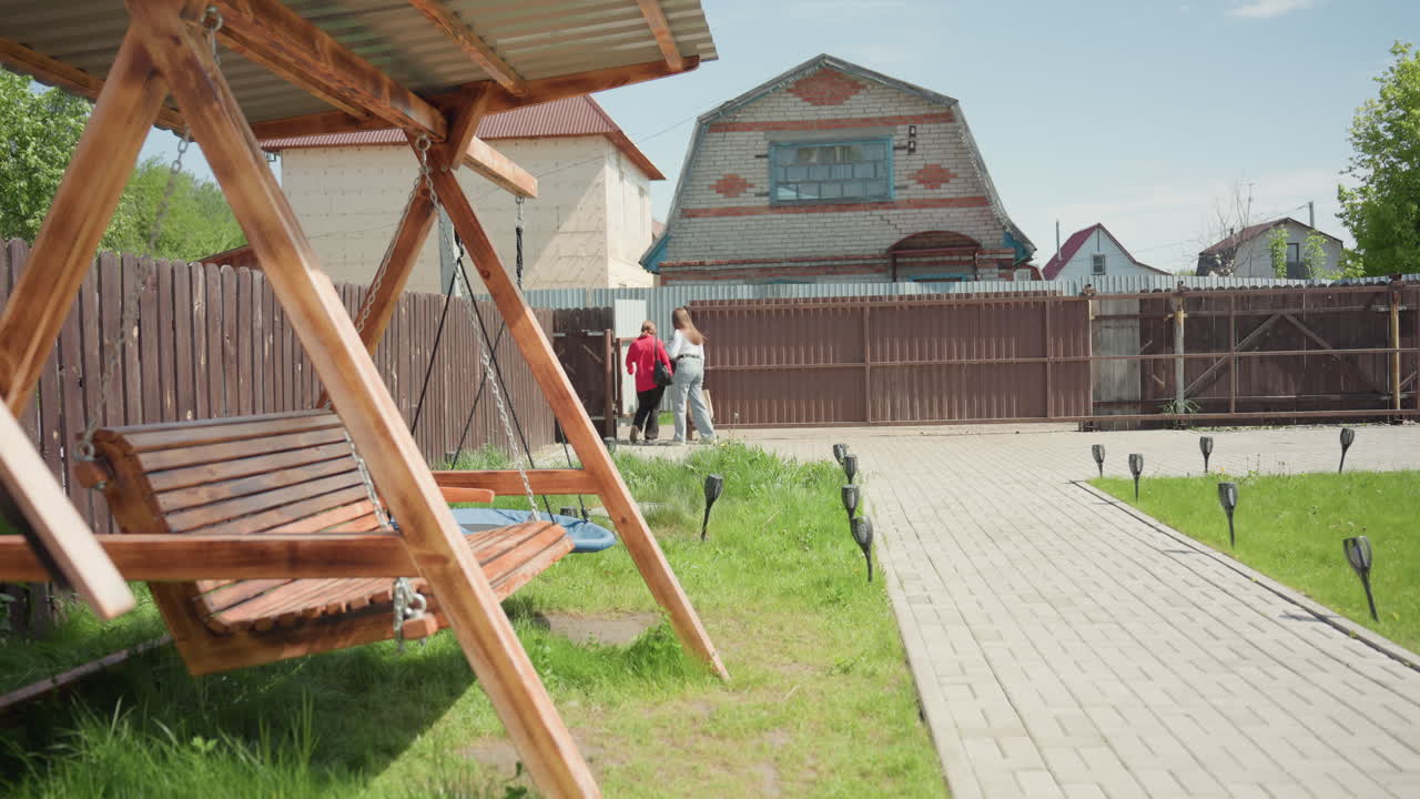 Three siblings casually walk out recreational centre along paved path, passing wooden swing under shelter on sunny day, surrounded by grass, wooden fence, and houses in quiet suburban neighborhood