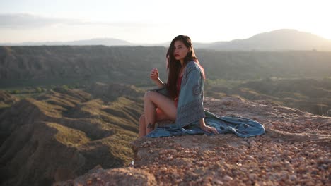 woman sits on a cliff overlooking a vast desert landscape at sunset