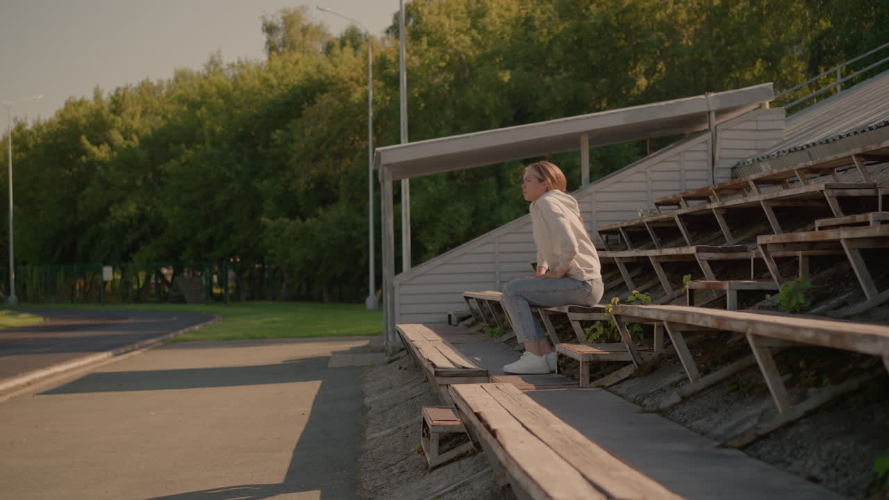 mujer en vaqueros casuales y sudadera con capucha camina por asientos rústicos del estadio con una postura relajada y reflexiva, la cabeza apoyada en la mano, el fondo incluye gradas vacías, postes eléctricos y árboles verdes vibrantes