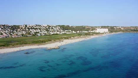 gente relajándose a lo largo de la playa de son bou en menorca, españa con aguas azules cristalinas y propiedades de lujo en el fondo