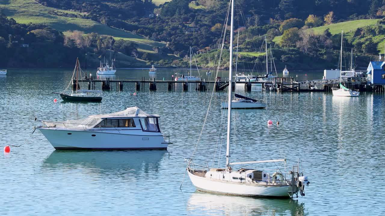 Boats gently sway in Akaroa Harbor under clear skies, capturing serene coastal scenery with calm waters and lush hills
