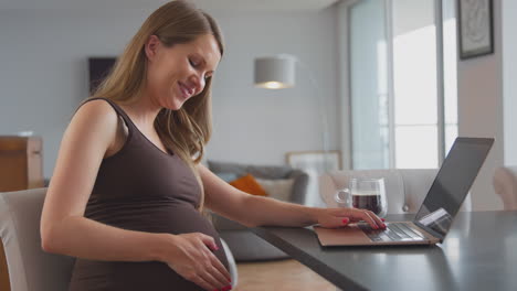 pregnant woman working from home on laptop sitting at table touching stomach