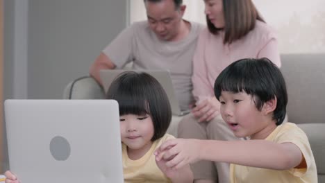 happy family, asian two young son with mom and dad sitting on sofa together. while using laptop computer browsing online internet watching cartoon, social media enjoy during holiday at home.