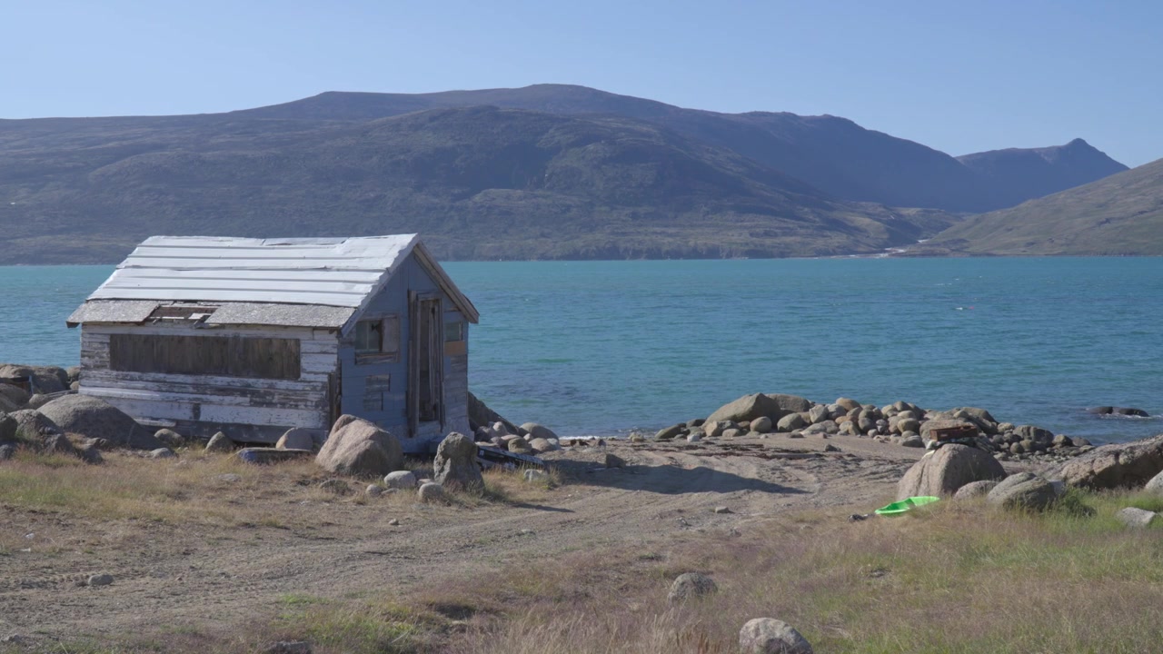 Vidvinkelbilde fra bakkenivå som fanger en liten, forvitret trehytte/utposthytte plassert langs den steinete kystlinjen med utsikt over solfylte turkise fjordfarvann.