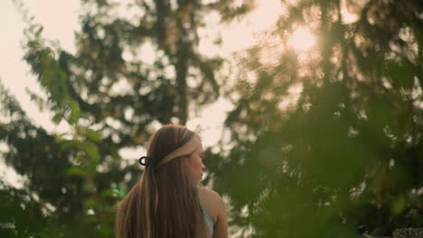 back view of young lady with hair tied back gazing thoughtfully to the right while sunlight filters through leaves, background blurred with soft bokeh effect from trees and distant lights