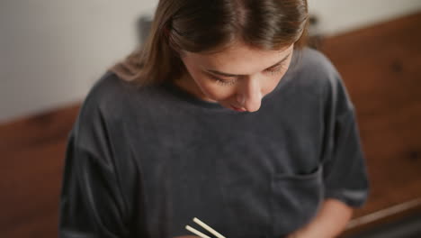 overhead close up of white lady wearing casual gray shirt eating meat with chopsticks, savoring delicious bite with focus and enjoyment, in bright kitchen with wooden countertop visible