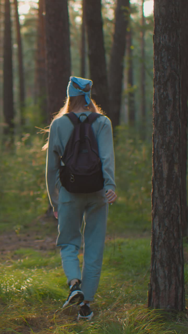 vista trasera de una mujer joven caminando a través de un bosque tranquilo, frotando juguetonamente su mano contra la corteza del árbol mientras lleva una mochila, la luz del sol se filtra a través de los árboles, proyectando un brillo cálido en su camino