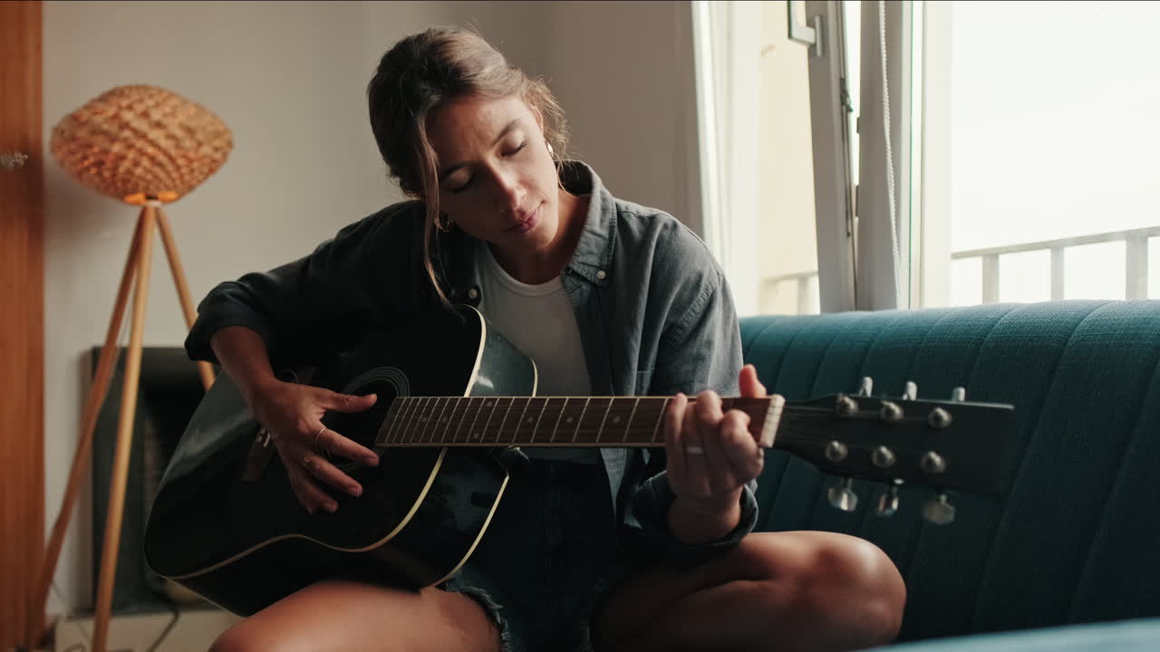 Ung kvinde spiller akustisk guitar på sofaen derhjemme