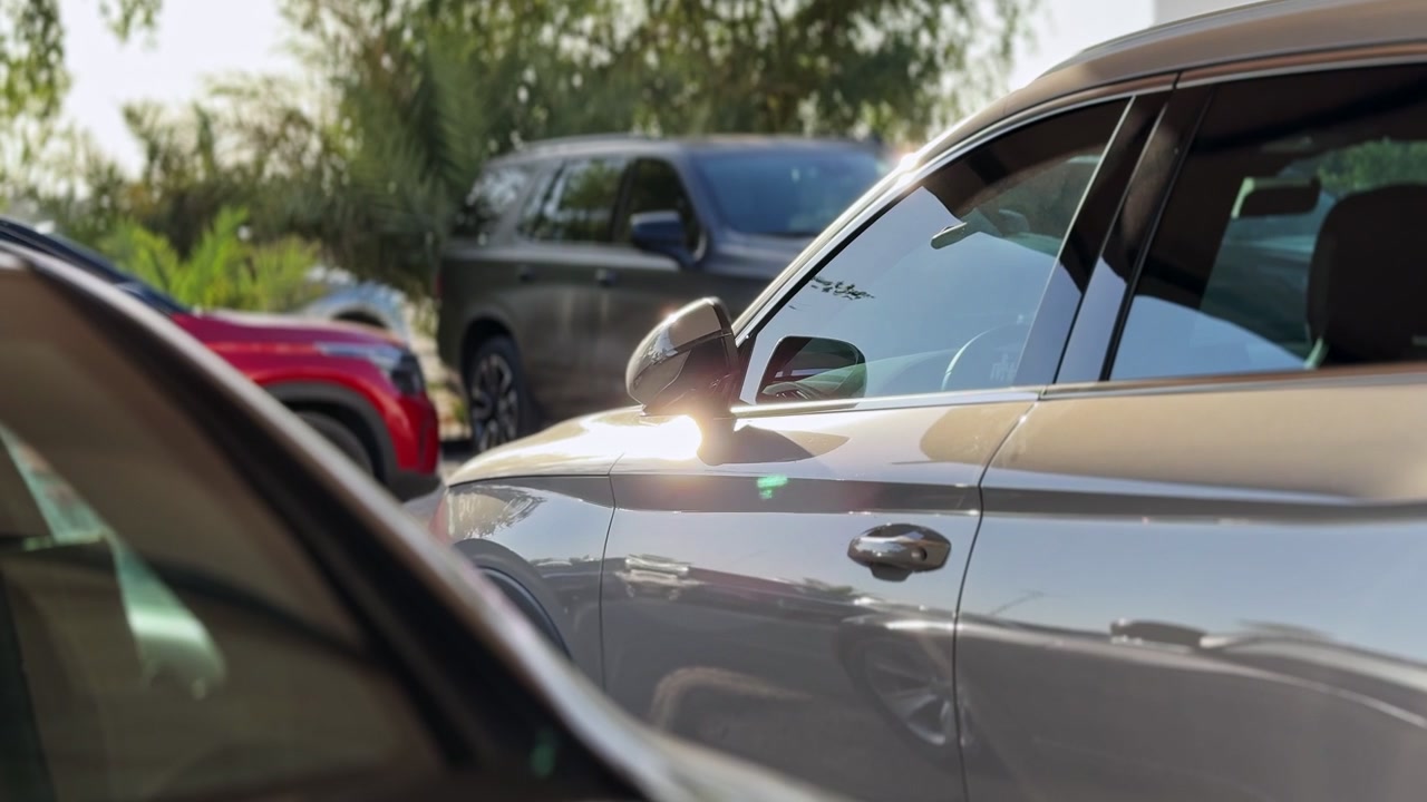 Vista lateral en primer plano de un coche plateado aparcado en un estacionamiento al aire libre en un día soleado. La luz solar brillante se refleja en la pintura metálica y el espejo lateral, con vehículos y árboles borrosos detrás.