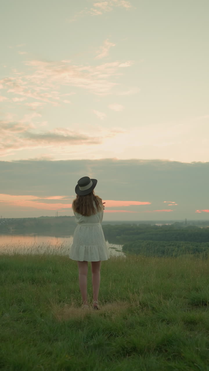 하 드레스와 모자를 입은 한 여인이 잔디에 서서 해가 지는 동안 조용한 호수를 바라보는 잔디 에 생각나게 서 있습니다. 그녀는 조용한 환경을 바라보며 평화롭고 반성적인 순간을 포착합니다.