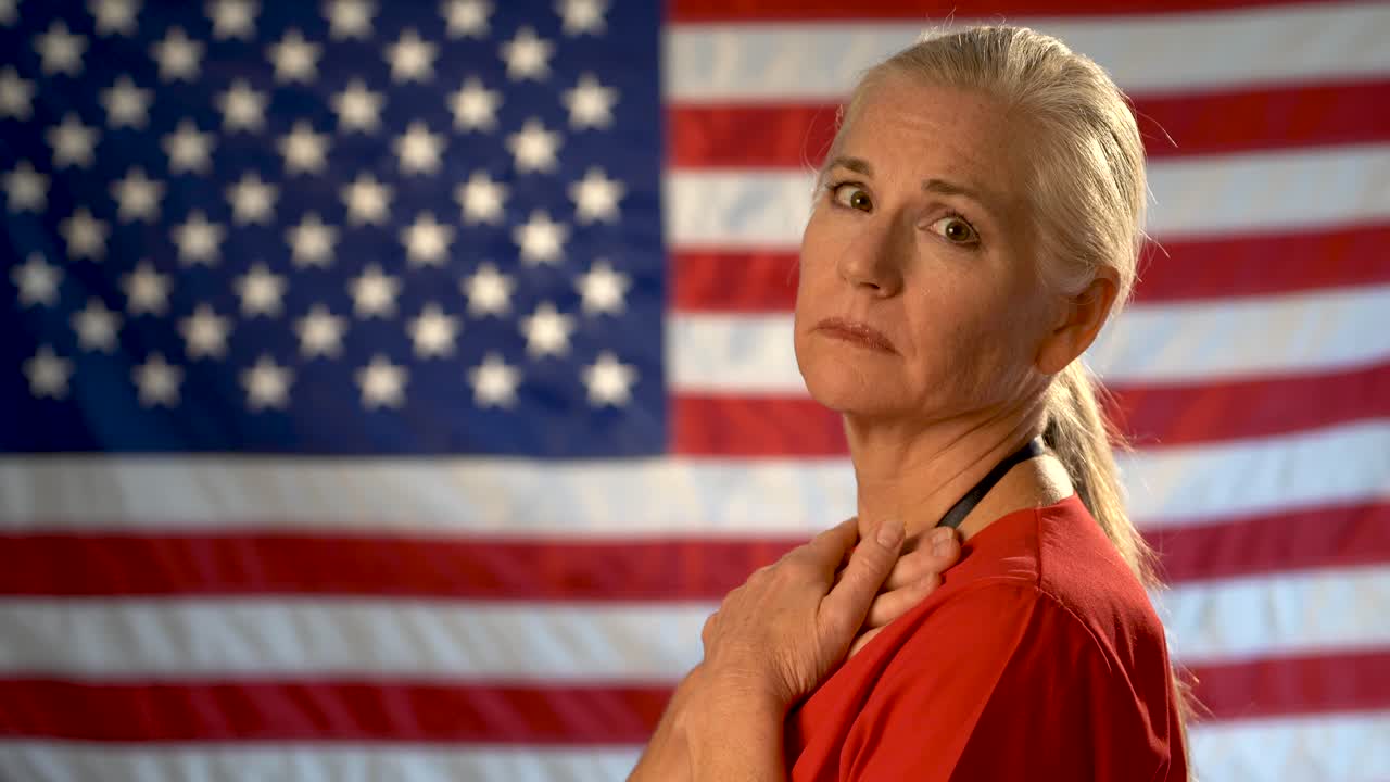Medium tight portrait of blonde nurse looking at camera appearing concerned and sad with out of focus US flag