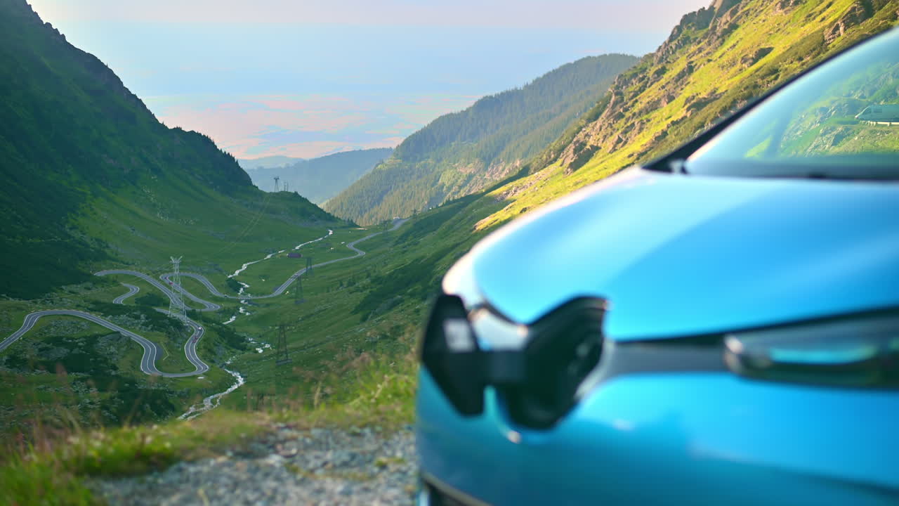 Uomo che collega un caricabatterie a un'auto elettrica sulla strada Transfăgărășan in Romania. Strada con pendii montuosi ricoperti di vegetazione sullo sfondo