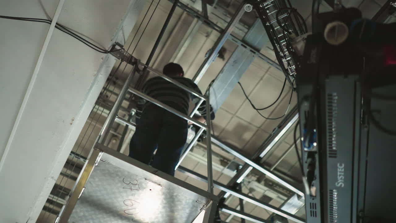 Arbejder klatrer op ad stige i industrielt kontrolrum, arbejder med udstyr og kabler, højvinkelbillede. Fokus på kabler og arbejderens position i hængende stillads.