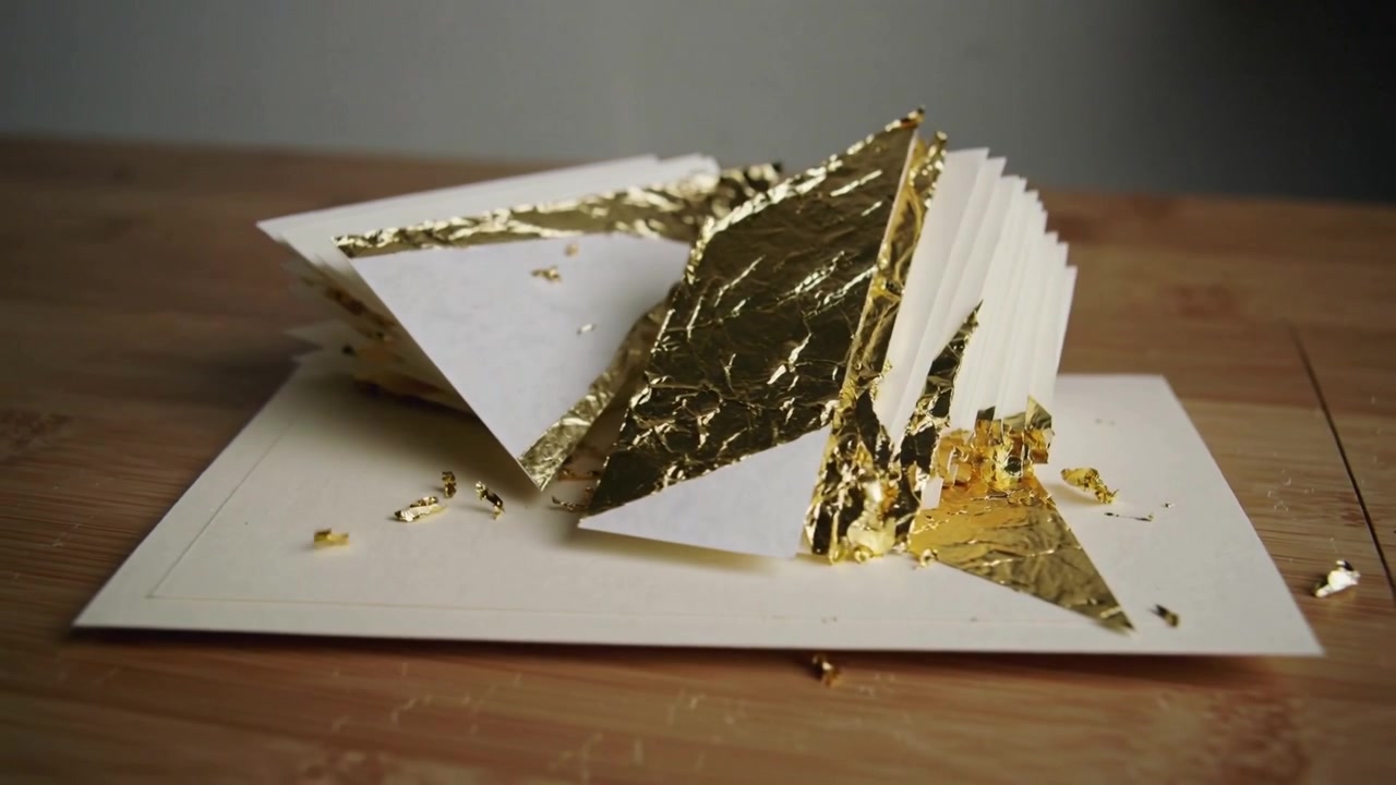 Telecamera che si sposta mostrando pieghe di carta che perdono scaglie di foglia d'oro mentre la lamina si solleva su un cartoncino crema, in studio.