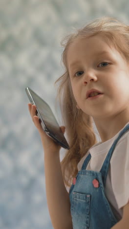 cu: adorable little girl with long ponytails in denim dress talks on smartphone spending time in light room of contemporary flat staying home closeup