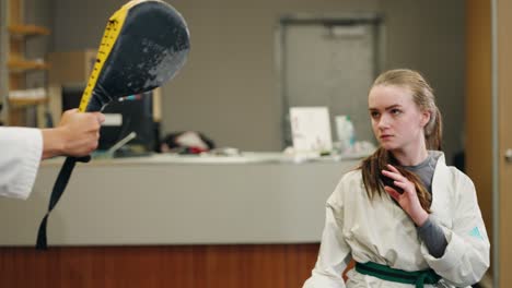 a young female combat fighter taekwondo karate martial arts student does slow motion practice training spinning close up high kick into pad with black belt master inside a exercise strength gym studio
