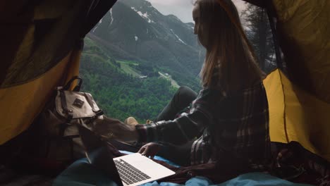 a girl hiker dressed in a plaid shirt sits in a yellow tent, uses a laptop. remote freelance work in the mountains