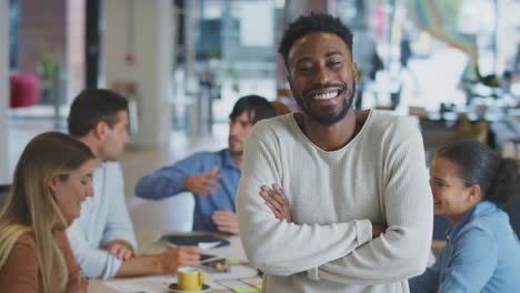 portrait of businessman with colleagues in background sitting around table in open plan office