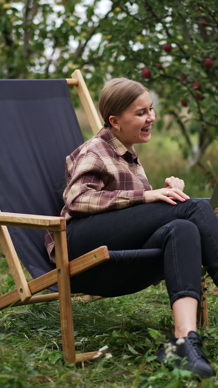 Caucasian woman watching video on laptop sitting outdoors. Lady is smiling happily and then looks astonished. Nature backdrop. Vertical video