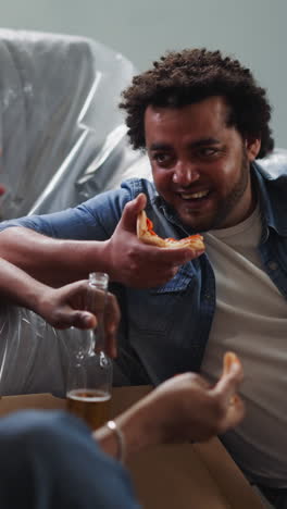 young african-american builders eat pizza with beer and tell jokes sitting near couch in renovated room. young black men celebrate repair finish in apartment
