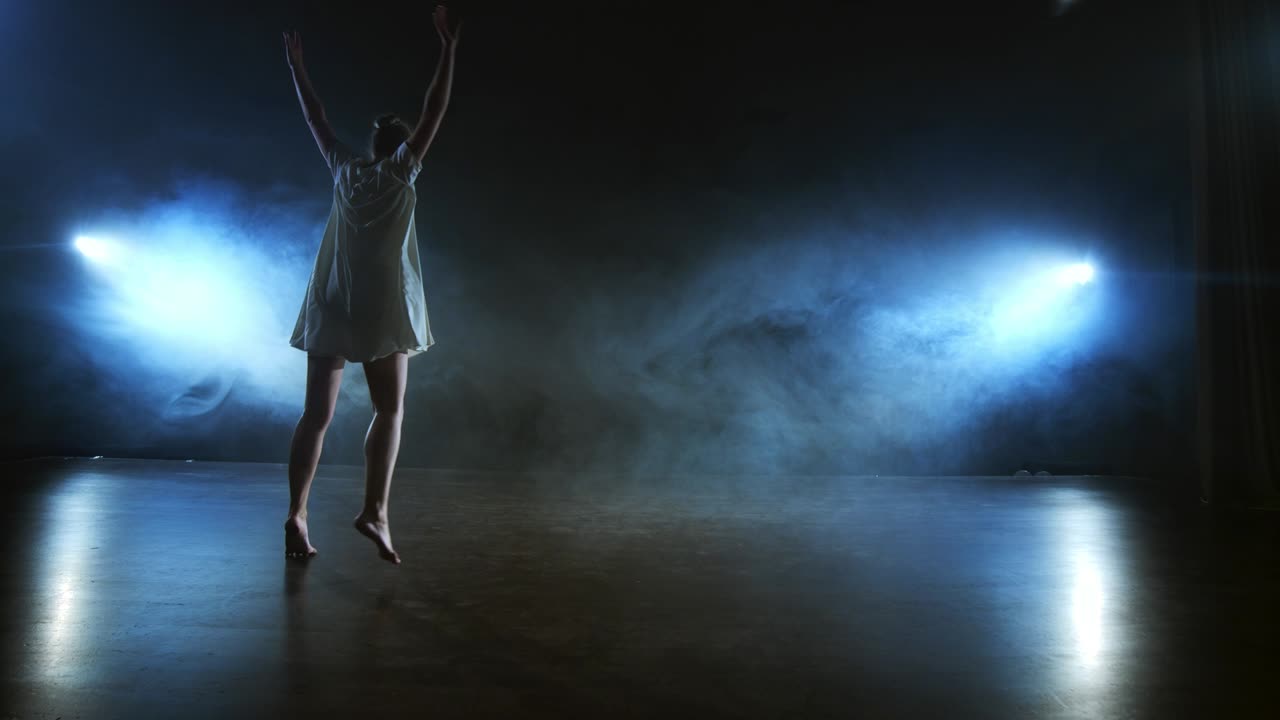 Modern dance woman in a white dress dances a modern ballet, jumps, makes rotation on the stage with smoke in the blue spotlights.