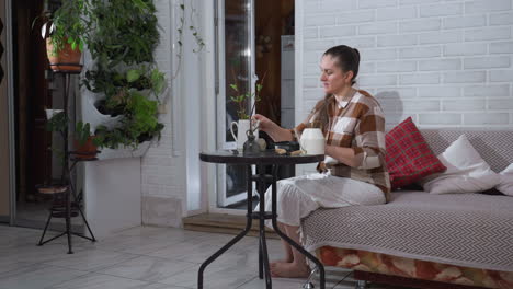 long shot of plant loving creative seated on sofa beside lush vertical garden gently sprinkling into plastic plate on round glass table surrounded by watering can, tools, indoor plants