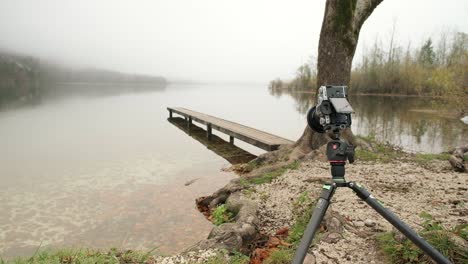 la cámara instalada en un trípode muy cerca del suelo en un día de niebla se acerca al borde de un lago con un muelle de madera en el fondo