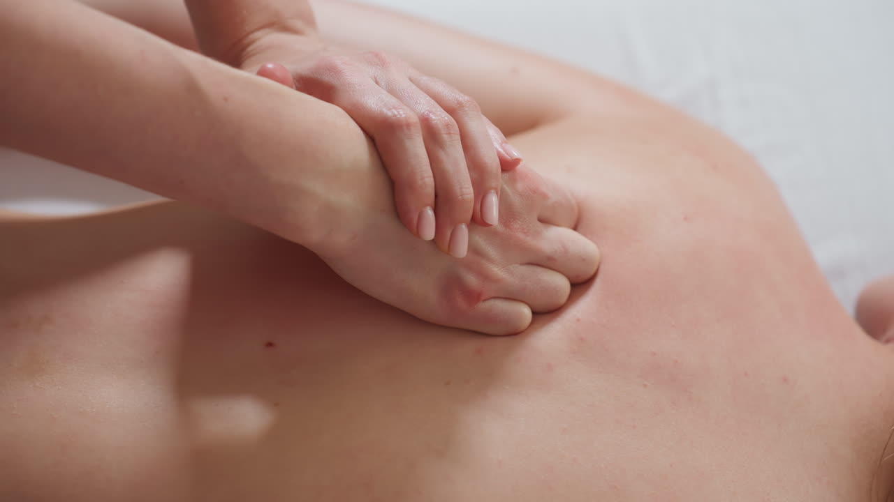 Hands of light skin woman performing gentle massage on back of client with light blemishes, captured in close-up view showing therapeutic motion, smooth touch, and serene wellness environment