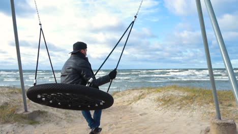 man watching baltic ocean sea waves moving in slow motion while sitting on a swing at a beach during autumn stormy and cloudy day. man wears a black leather jacket and dark clouds are in the sky.