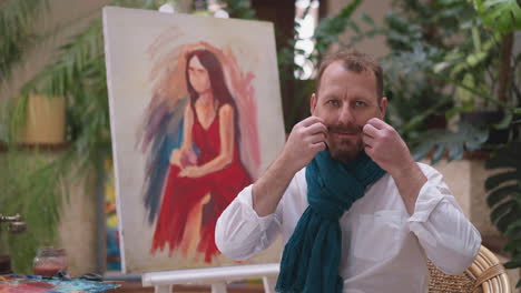 man in modern scarf fixes mustache near portrait. artist creates visually intriguing juxtaposition between personal grooming and act of artistic creation