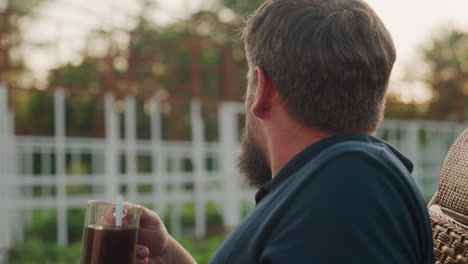 side profile of young bearded man seated in wicker rocker outdoors sipping steaming black coffee slowly enjoying tranquil sunset garden atmosphere warm light calm reflective moment