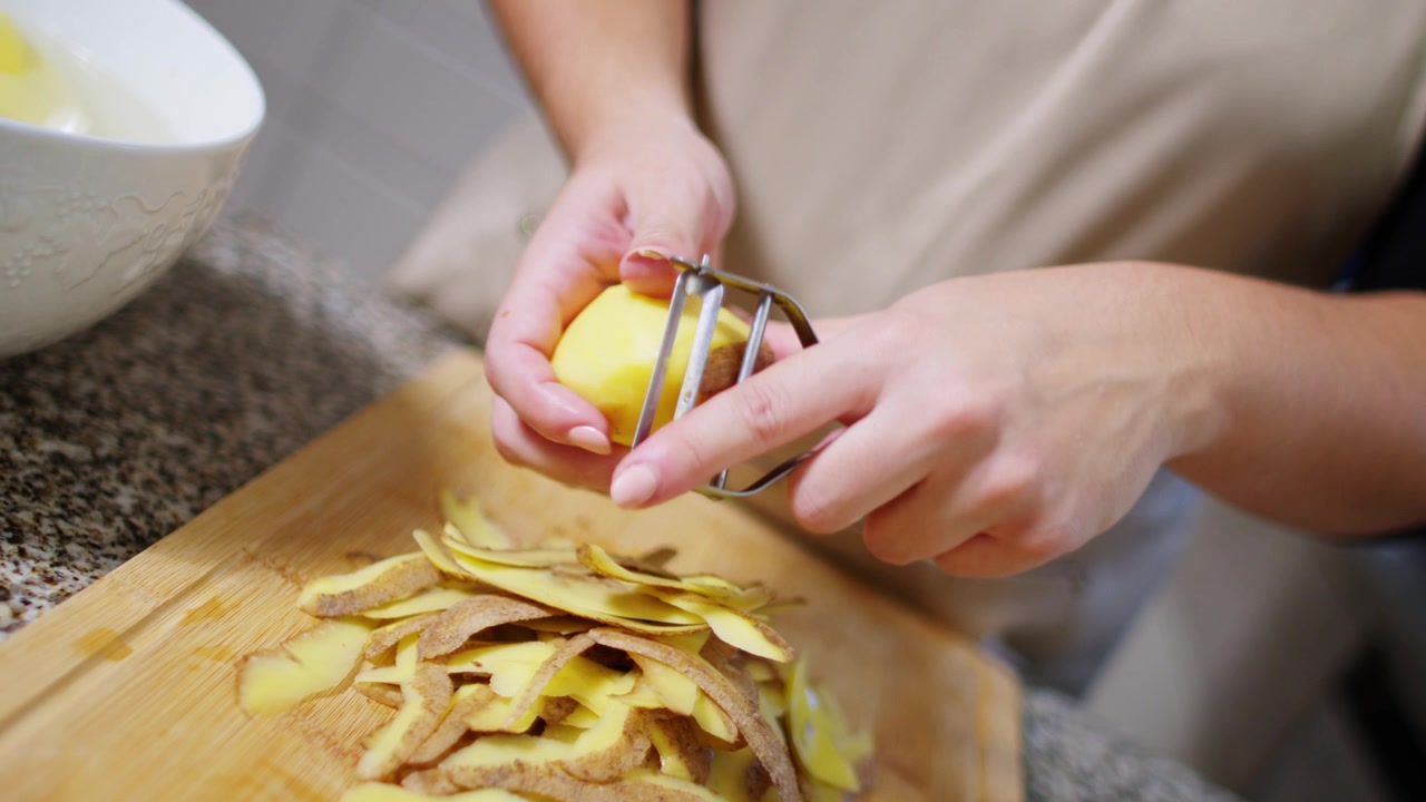 Filmato al rallentatore di una donna che pela patate con un pelapatate, con le bucce che cadono su un tagliere di legno.