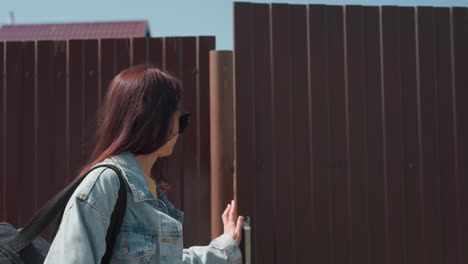 two young women walk through brown metal door into sunny outdoor area, first girl in red shirt leads with neutral expression while second woman in sunglasses follows close behind