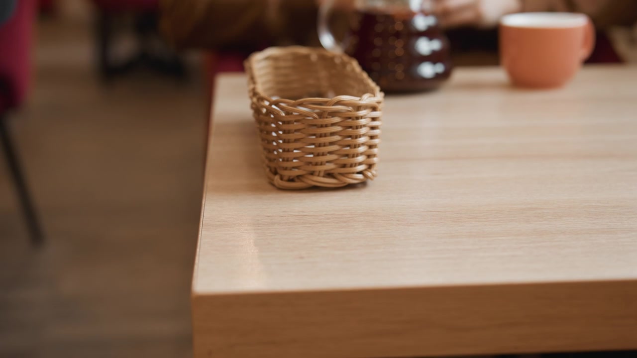 Close up of hands placing small wicker basket on wooden table. Background shows coffee pot and cup soft lighting in indoor setting with blurred background
