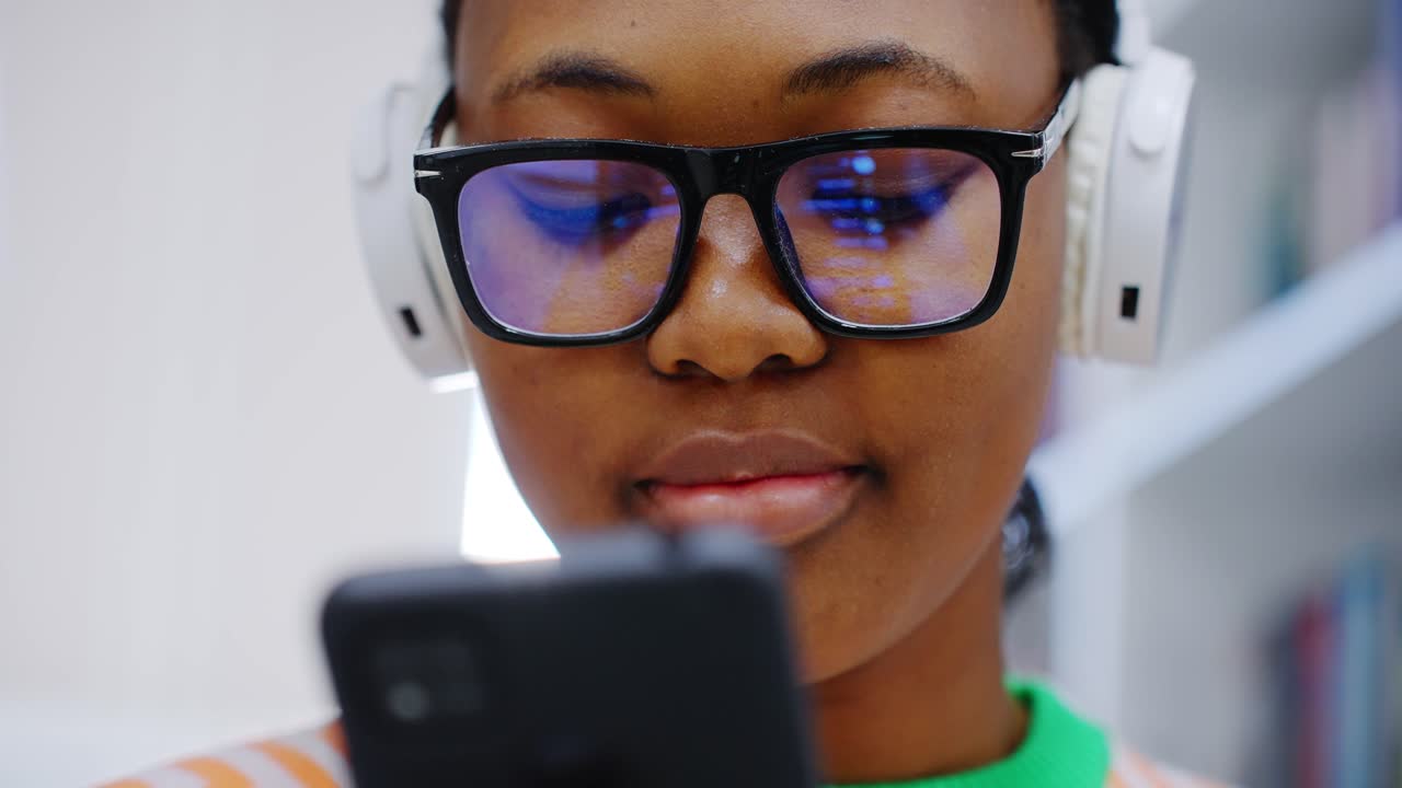 Koncentreret ung sort kvinde med briller og hovedtelefoner, der bruger telefon