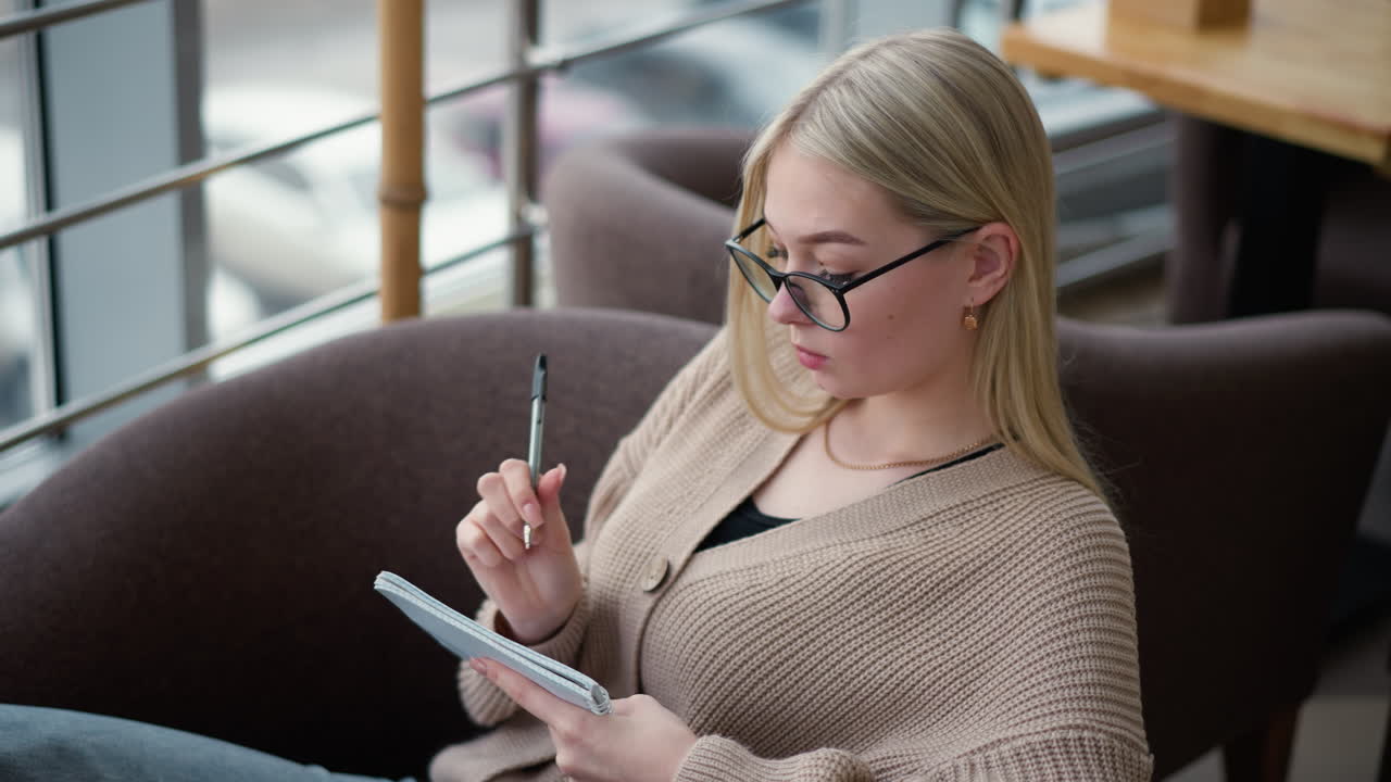 Student thoughtfully writing in notebook, focused on taking notes, she is in a study space with a calm and quiet atmosphere, wearing glasses and a cozy sweater