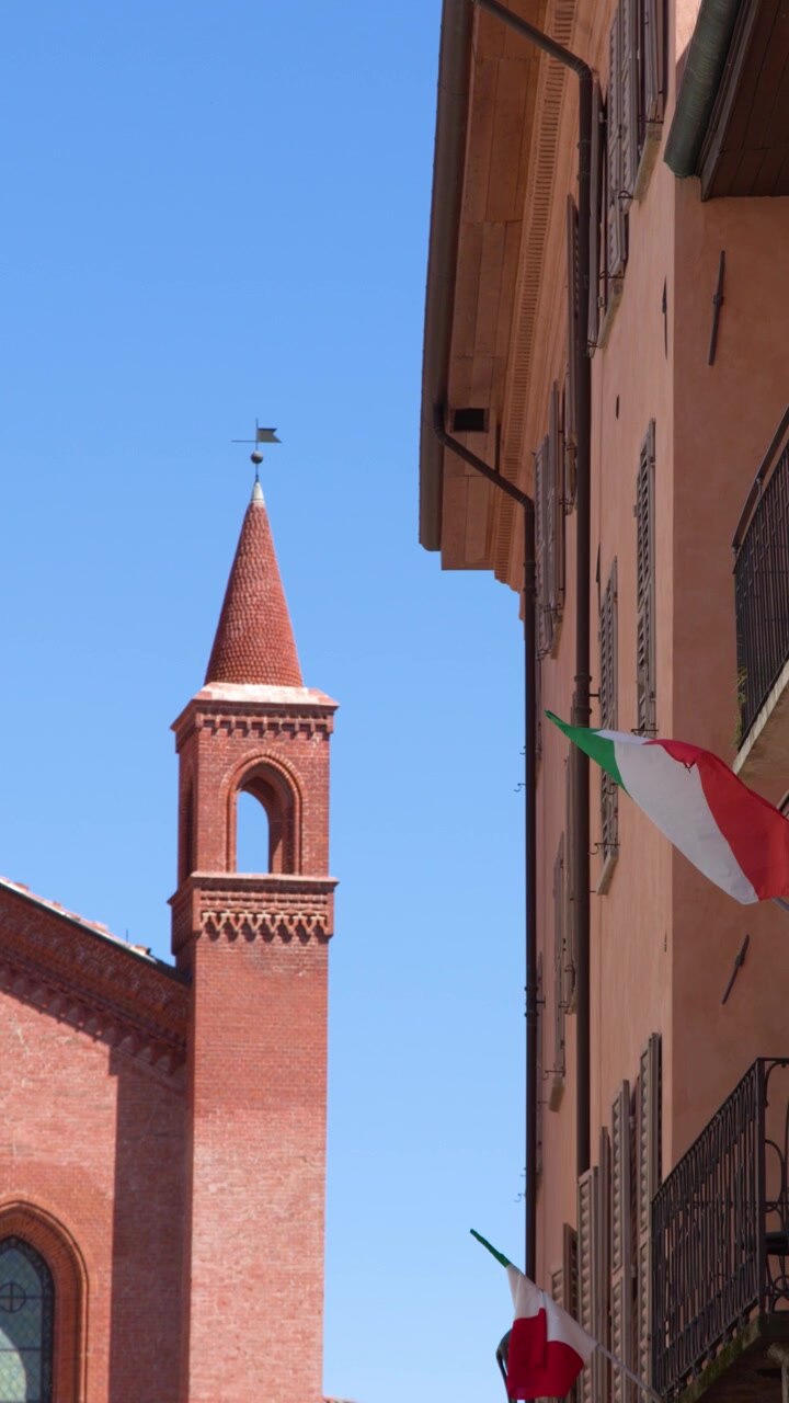 Vista dal basso tra edifici storici verso una torre cattedrale in mattoni e bandiere italiane che sventolano ad Alba