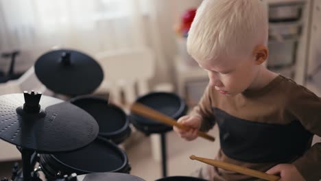 Close-up-shot-of-a-happy-albino-boy-with-white-hair-playing-a-black-electronic-drum-kit-using-special-wooden-sticks-in-his-room-during-a-day-off-developing-his-hobby-and-talent