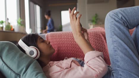 video of relaxed biracial woman with headphones lying on sofa and using smartphone