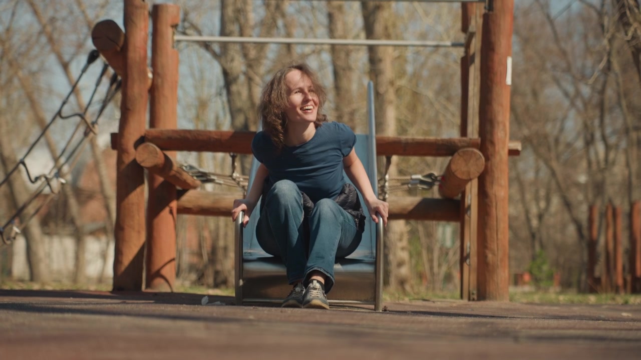 Close up of woman sliding down and sitting on playground equipment in outdoor park setting. Woman wears casual clothing including jeans sneakers and raises her arms during slide before