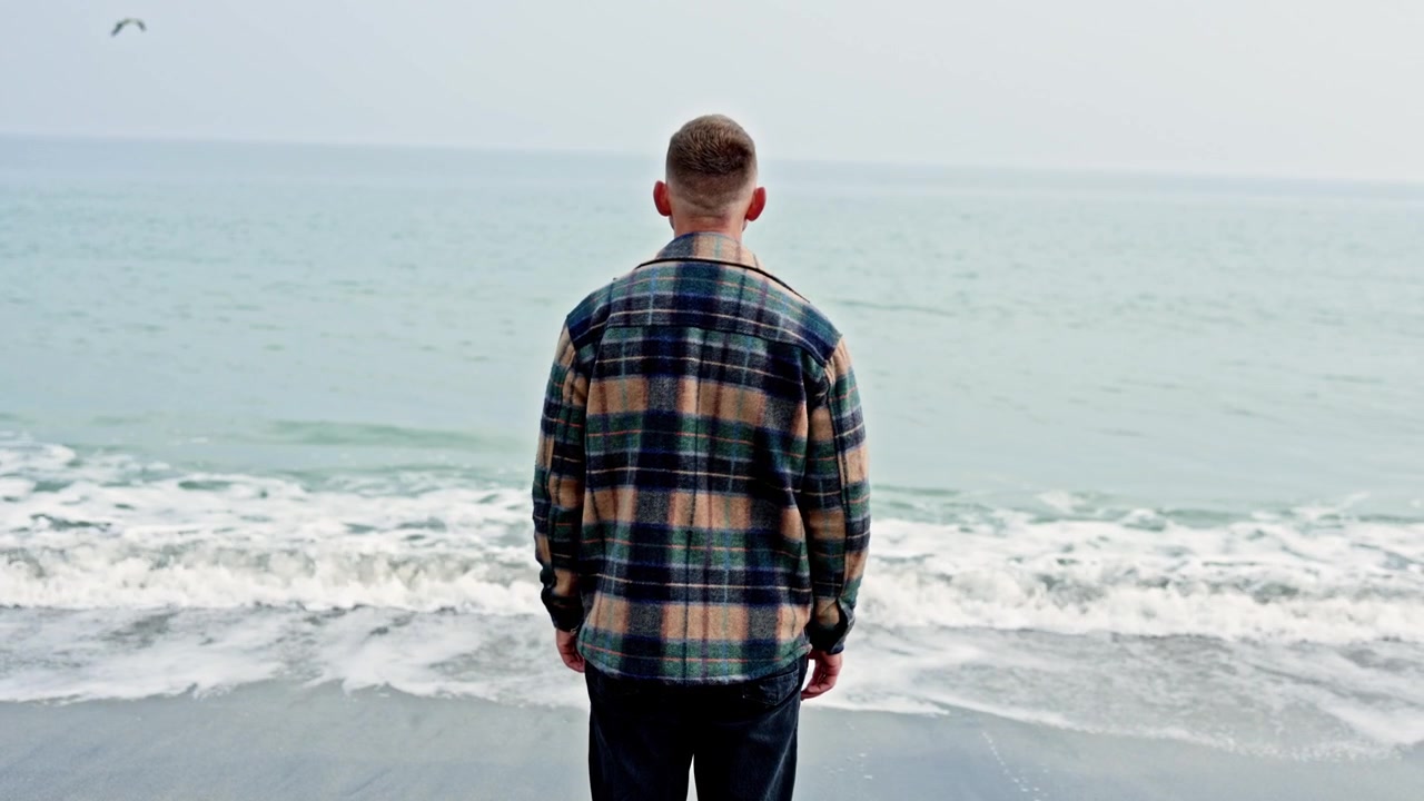 A young man stands on the shore and looks into the sea. A man walks by the sea in autumn. A man in a coat stands and looks into the sea.