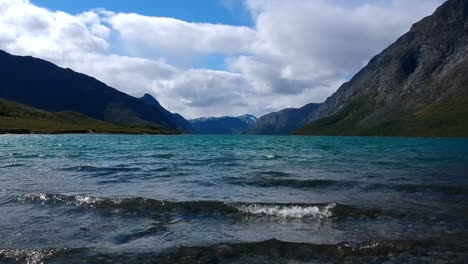 tranquil scene of a turquoise lake rippling gently against the shore, surrounded by majestic, dark mountains under a cloudy sky.