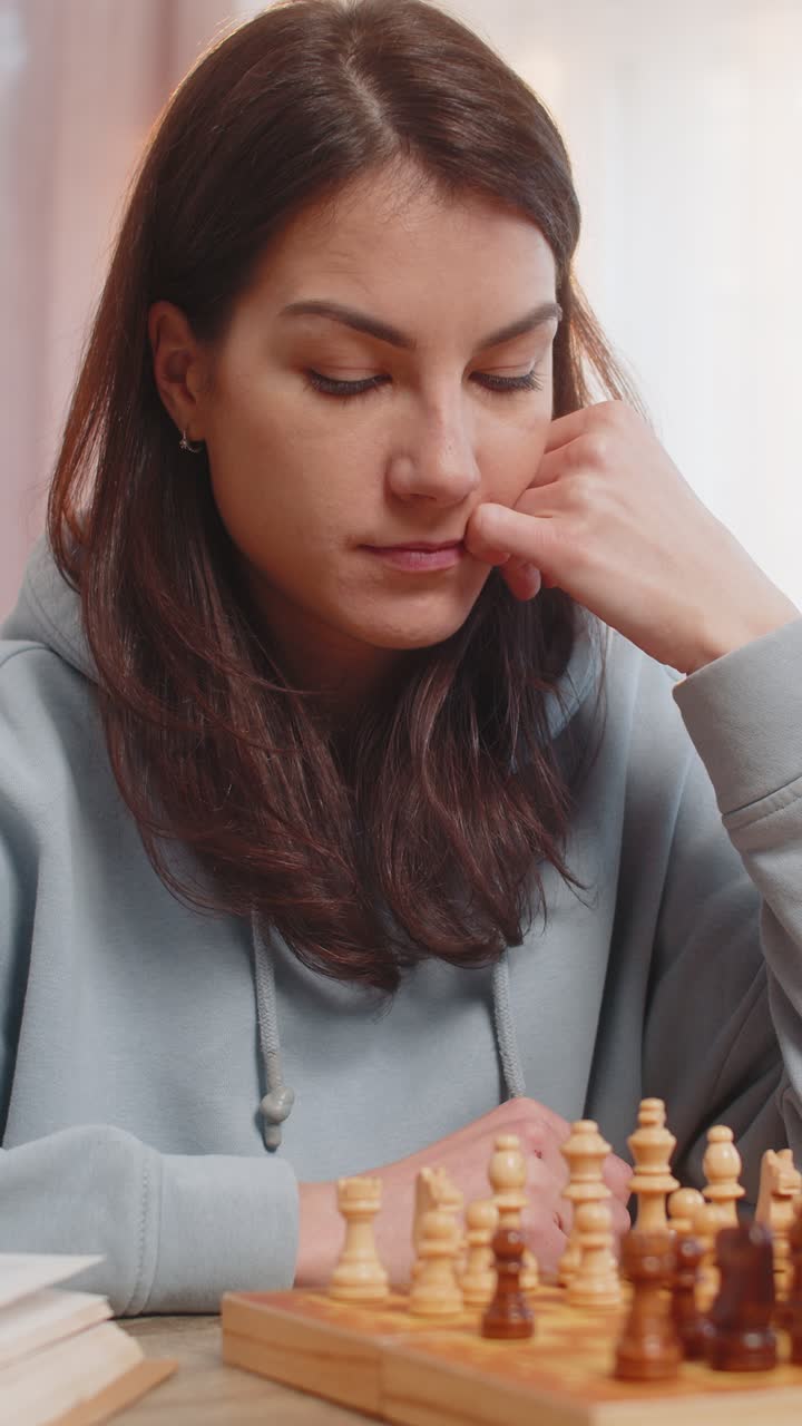 joven jugando al ajedrez sola arreglando piezas en la mesa en el sofá con el libro acostado cerca en casa
