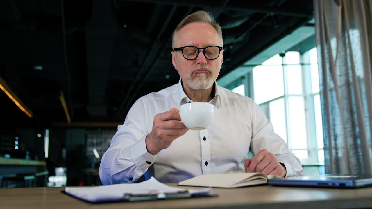 Positive adult Caucasian man sits at desk holding a cup. Man enjoying coffee at work and looking at window.